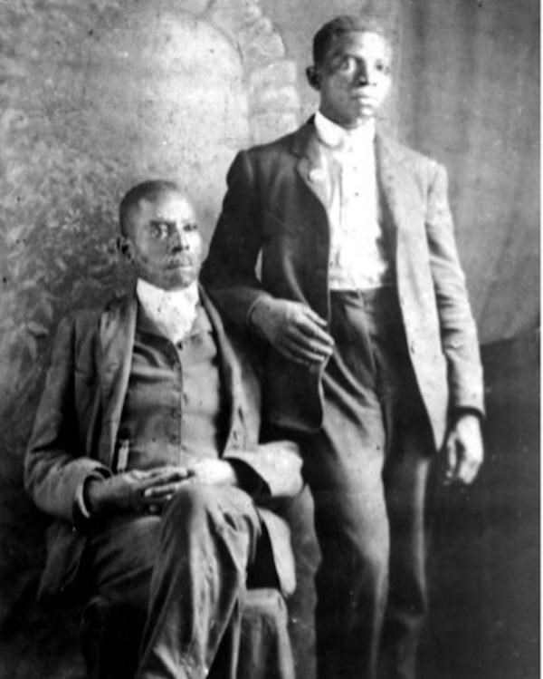 Black and white photo of older man sitting wearing a nice suit, his son stands next to him.