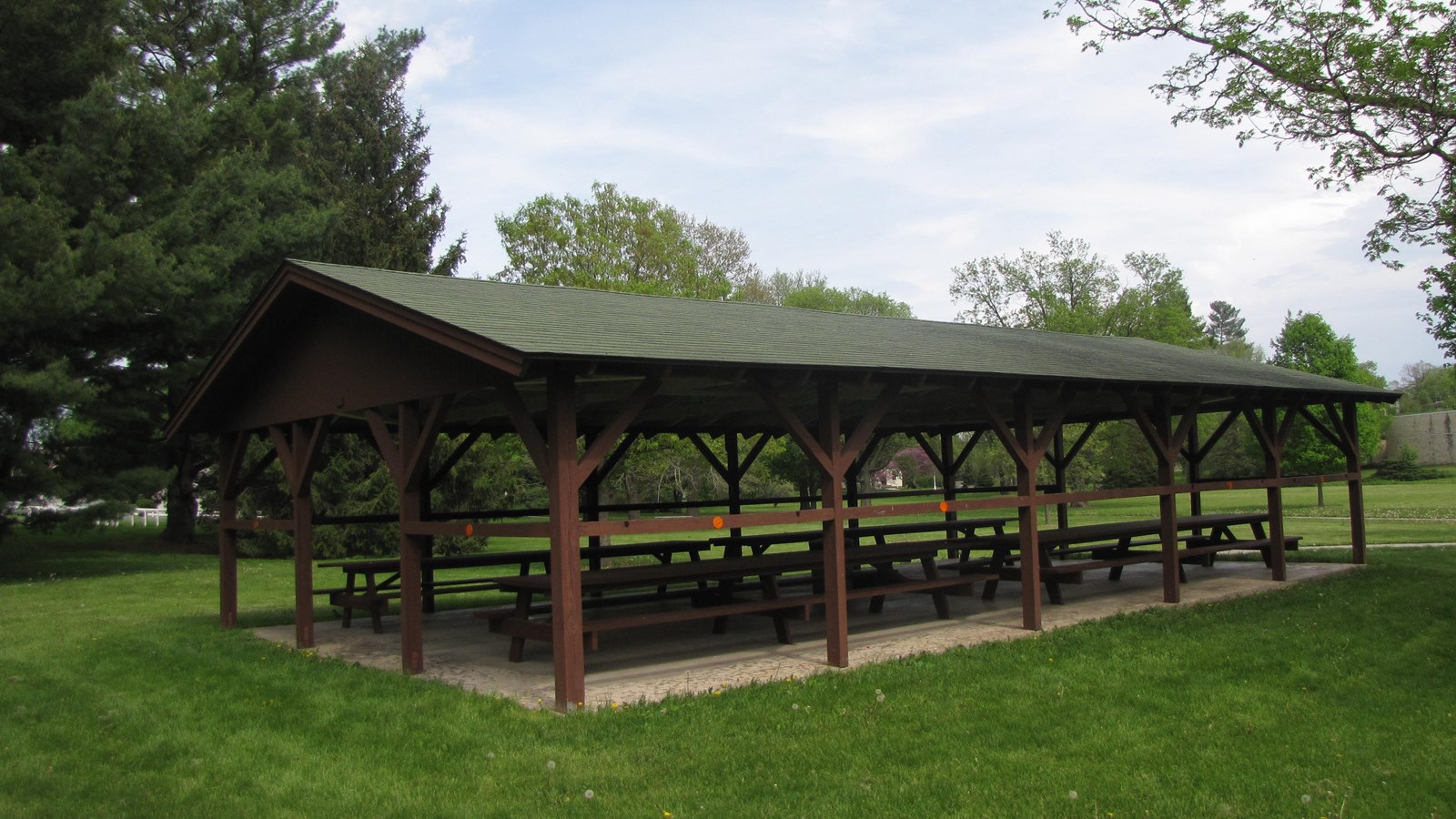 A park picnic shelter has a reddish-brown timber frame and green shingled roof.