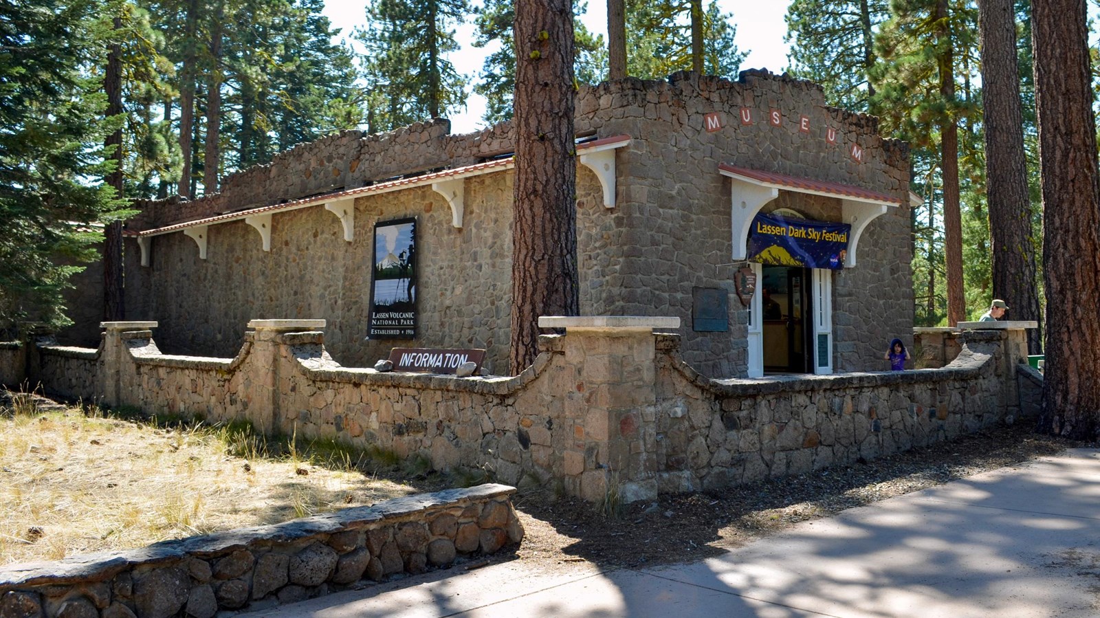 A one-story stone building surrounding by a 3-foot-high stone wall amid conifers.