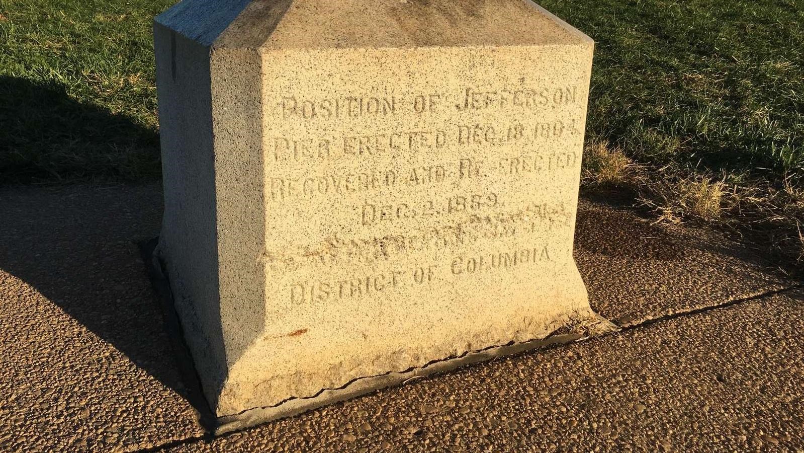 A stone block with words carved into the face on a concrete pad, surrounded by green grass.