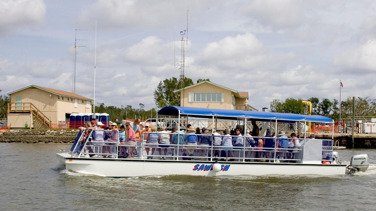 An open air white boat with a blue canopy is full of people. Shipping docks are in the distance