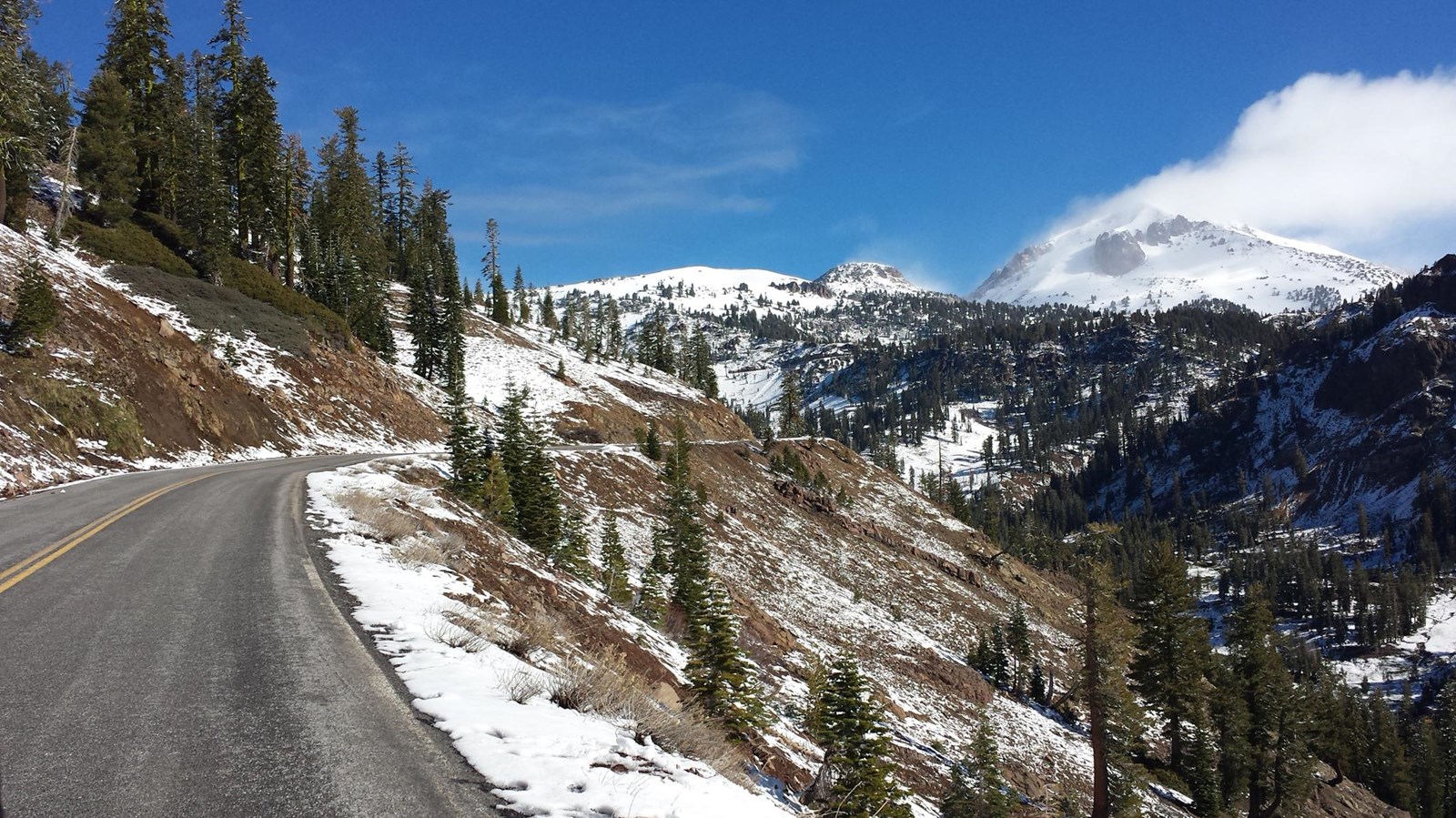 A photo of a highway crossing a mountain slope dotted with light snow. Clouds are lifting to reveal 