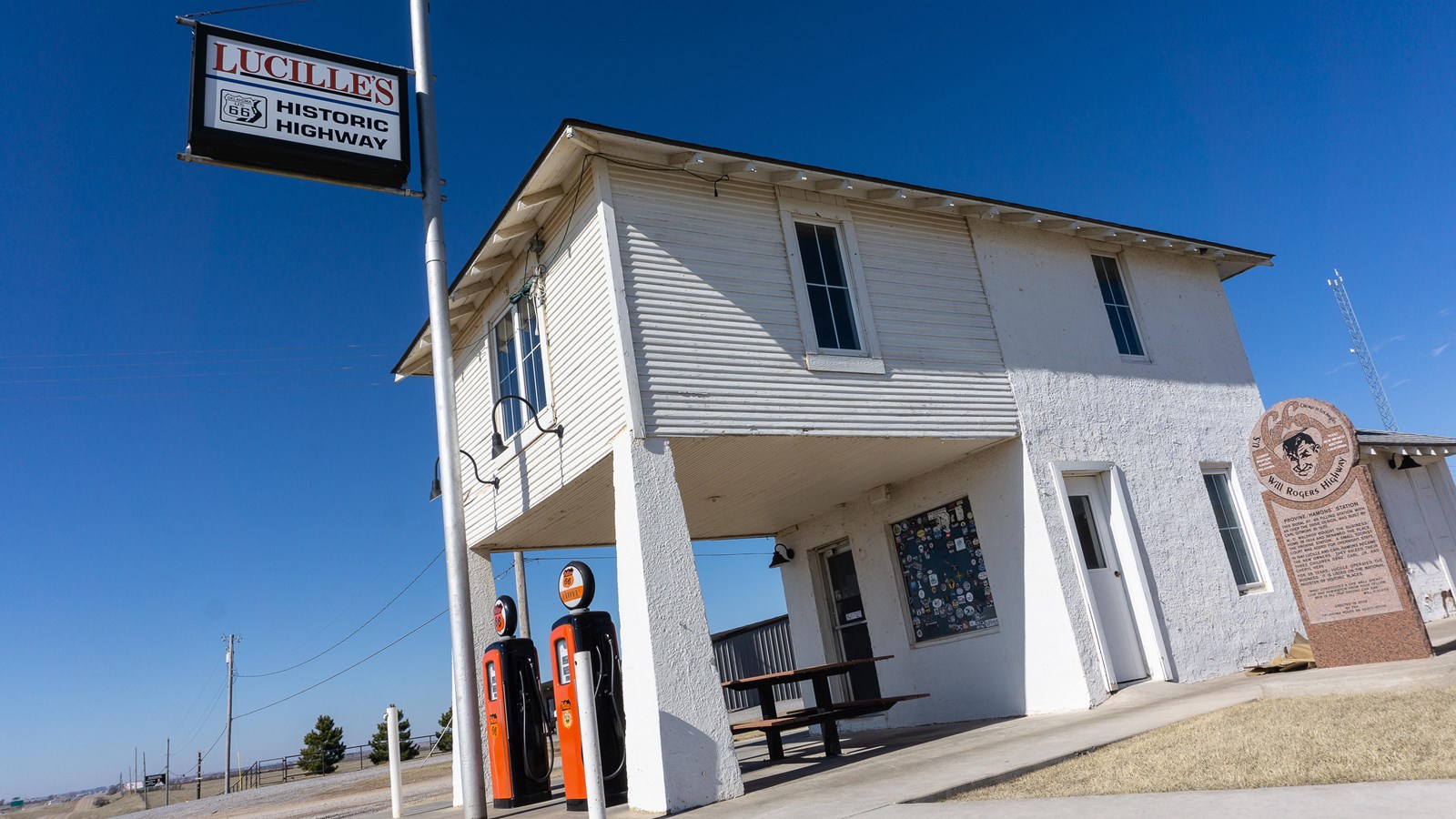 A white two story building with a covered patio and two old gas pumps. A tall sign reads 