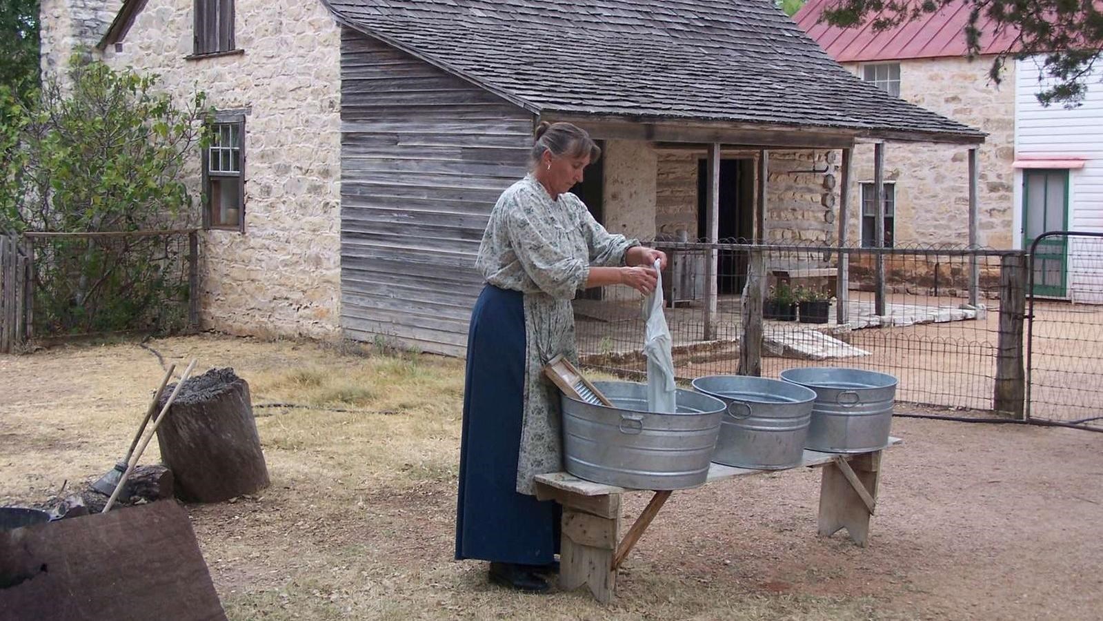 A woman does laundry outside in the yard of the farmhouse using a scrub board and metal washtubs.