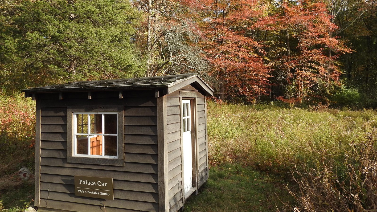 A small wooden house in a field with trees in the background with fall leaves.