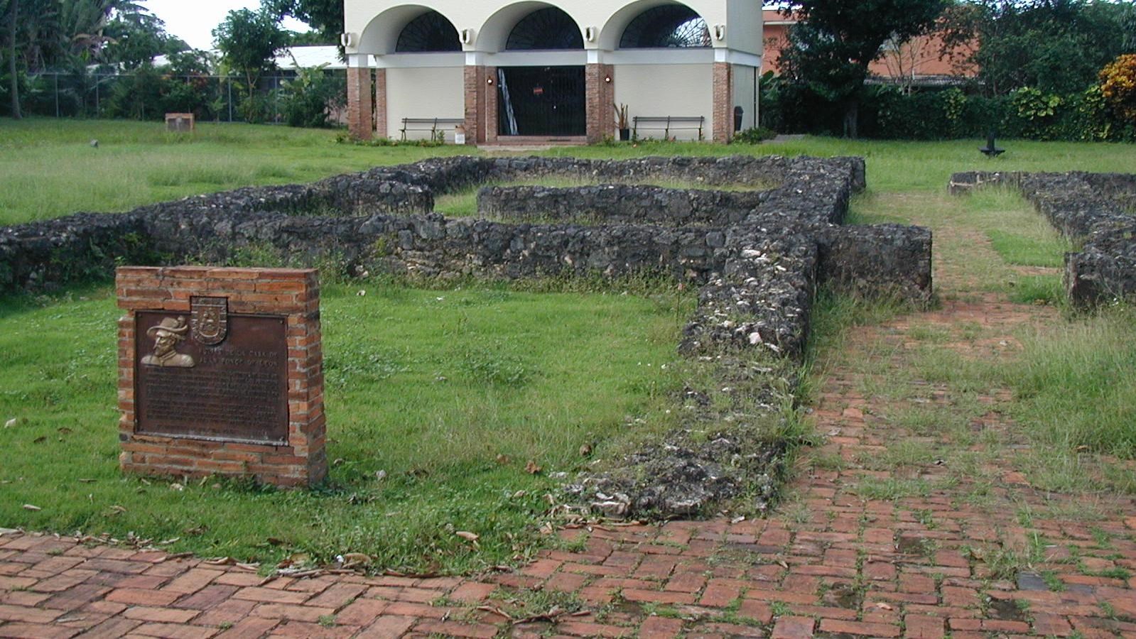 Dark stone foundation of buildings with a metal sign mounted on a brick wall in front of them.