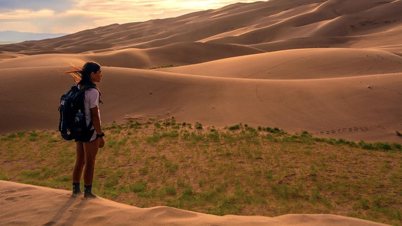 Girl with backpack standing on dunes at sunset