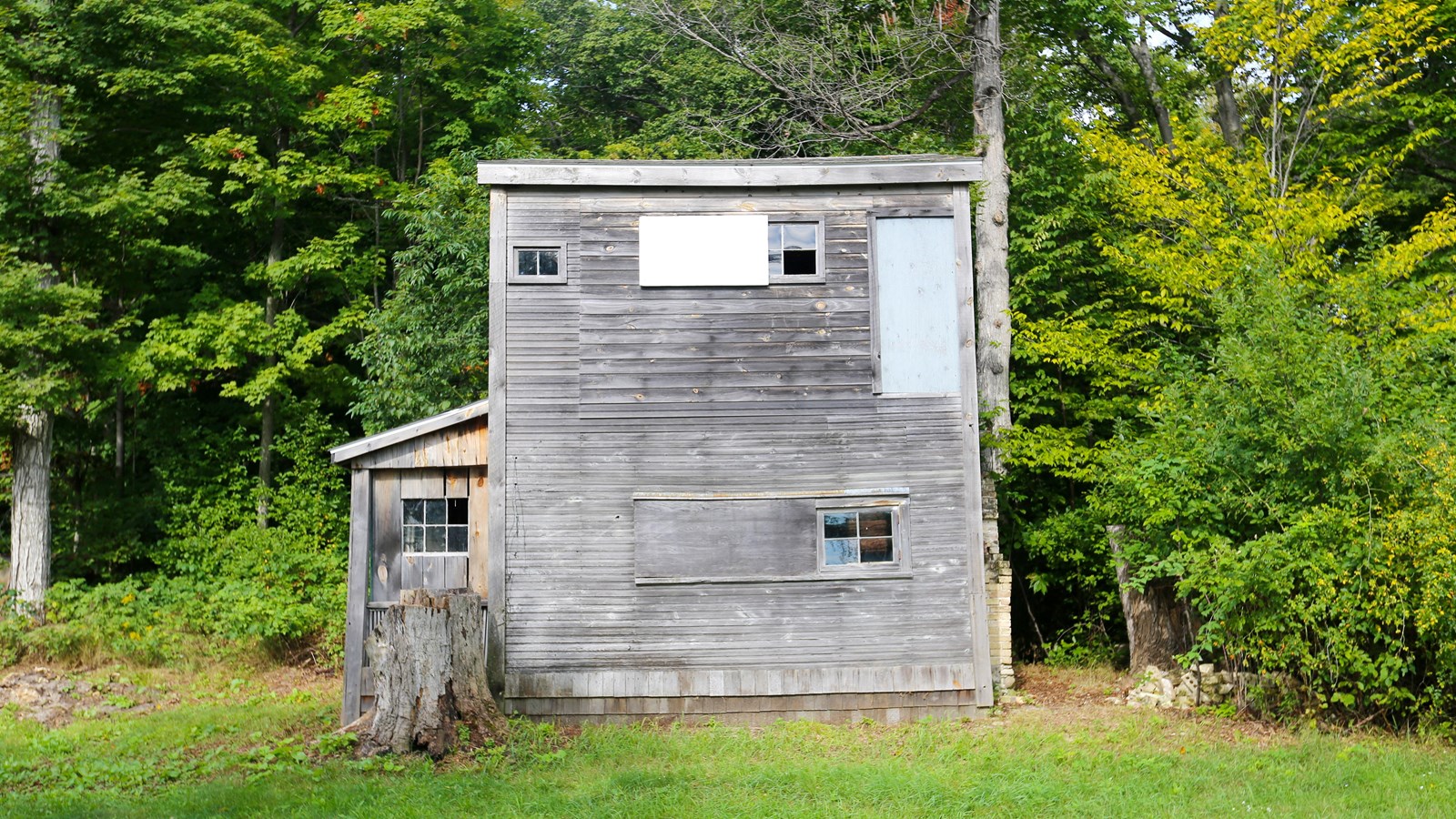Backed by woods, a 2-story weathered gray clapboard structure with a side-facing gable roof