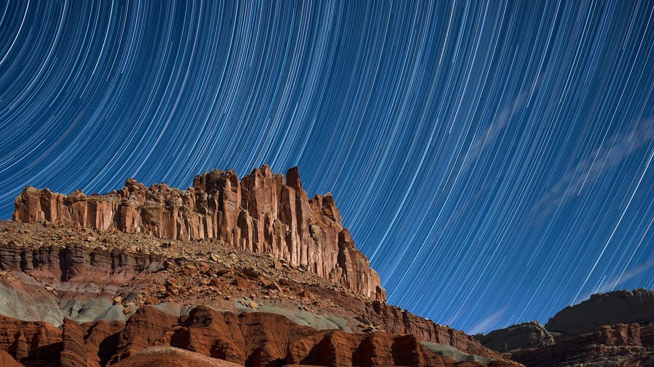 White lines representing circular star trails in dark blue sky, above colorful red cliffs. 