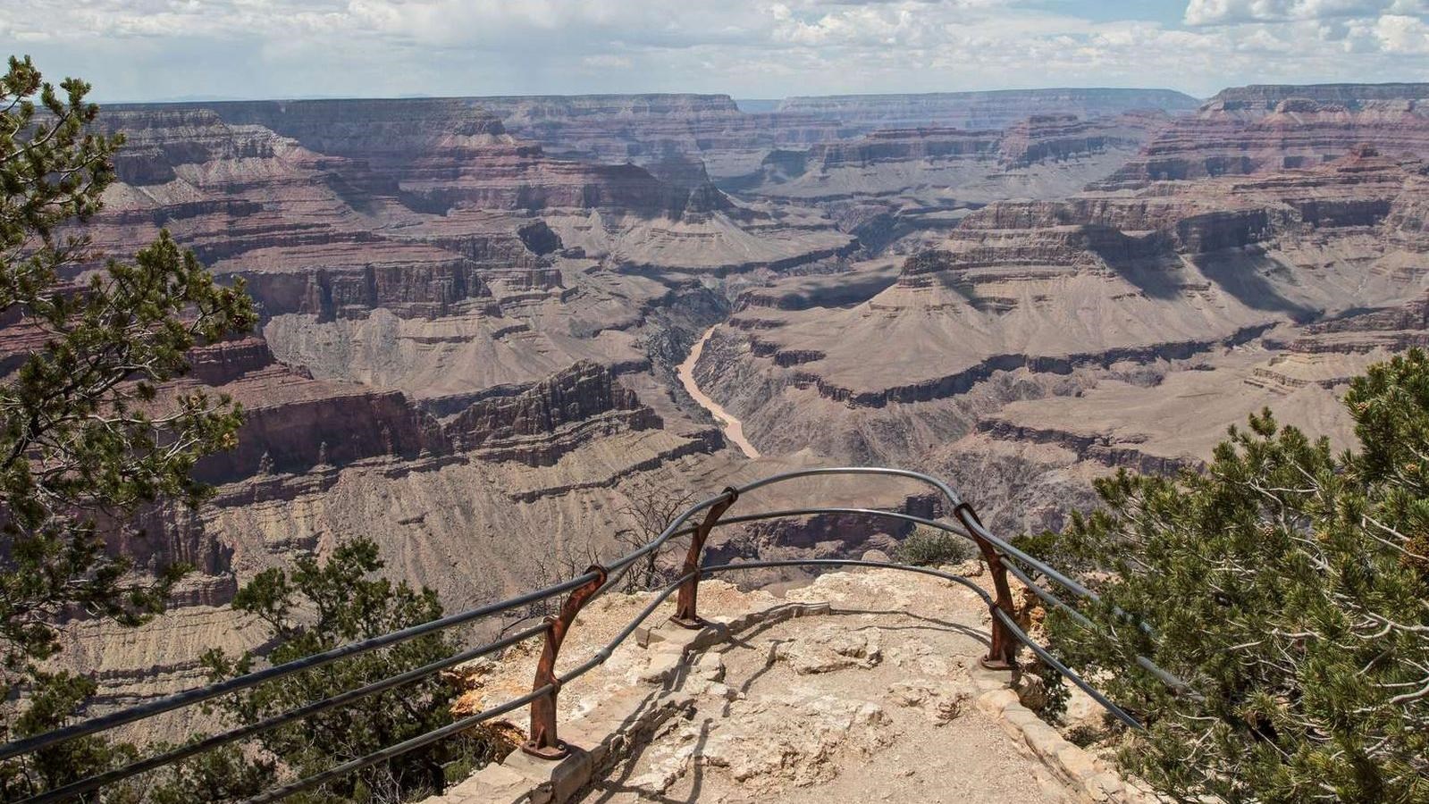 From behind a curved guardrail, a view of a river within a mile-deep canyon of peaks and cliffs.