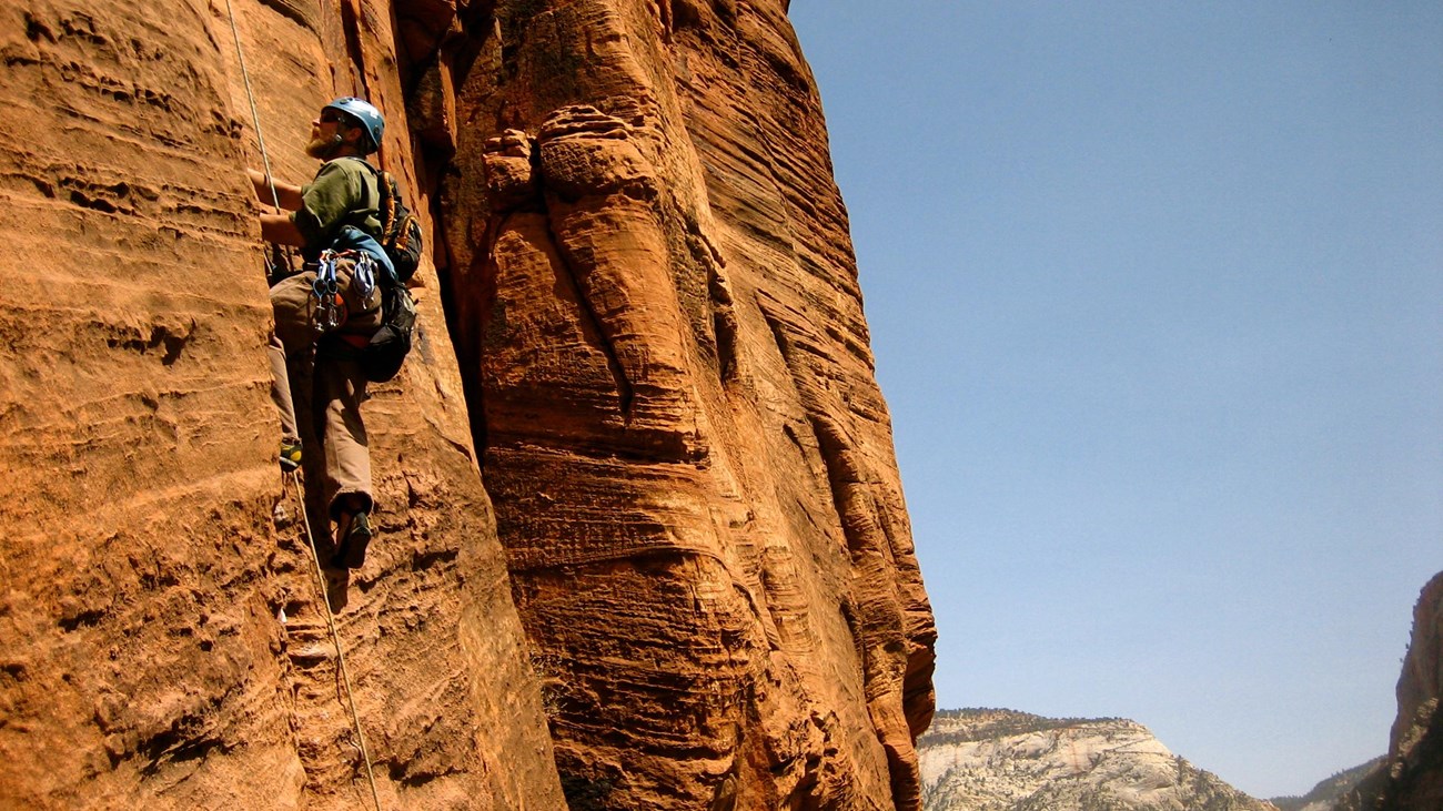Red-orange sandstone cliff-face with a rock climber in a helmet making his way up.