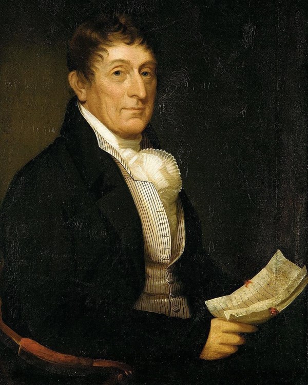 A man posed with cropped hair, a high collar vest with thin stripes, and holding a papers.