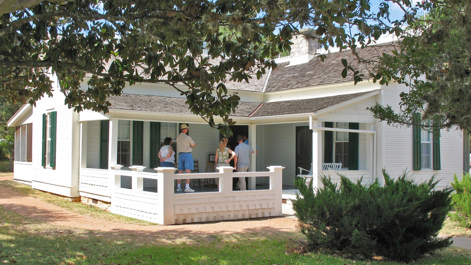 Visitors stand on the porch of a white house as viewed through a magnolia tree.