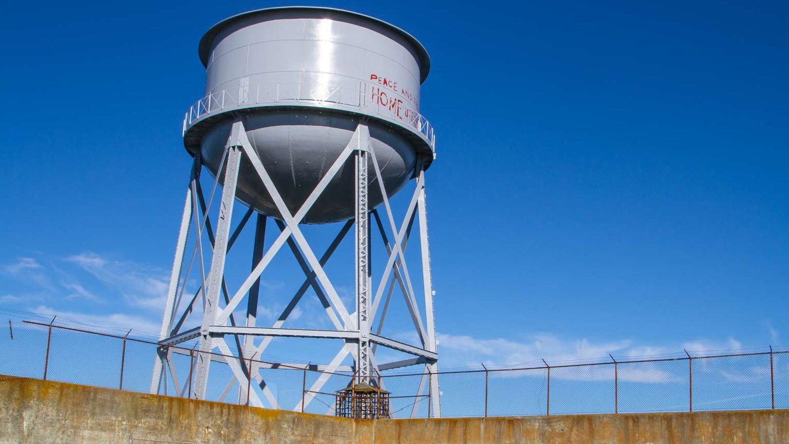 A view of the water tower on its scaffolding support looming large over a wall.