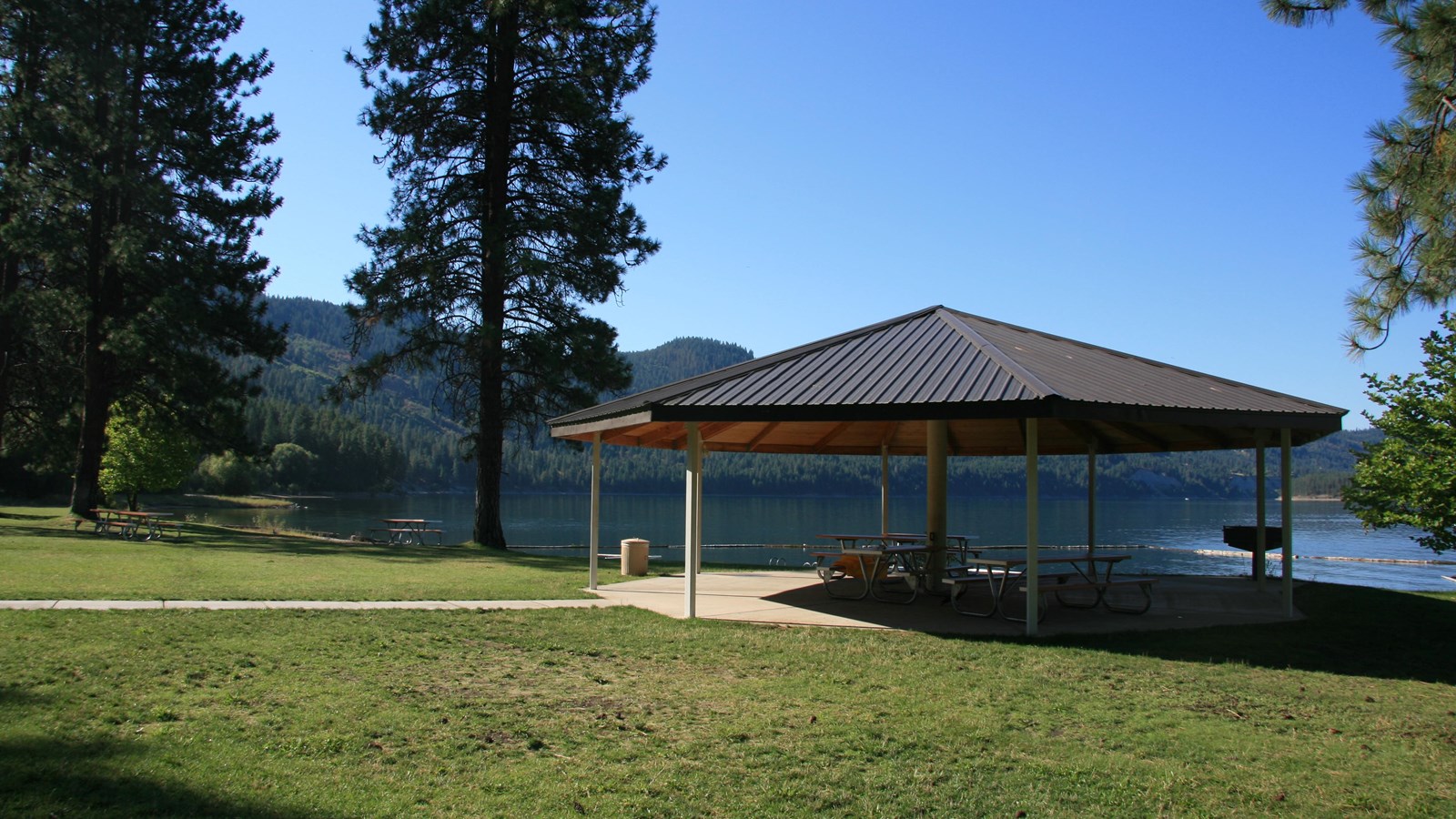 A roofed gazebo covers picnic tables and a grill adjacent to the lake.