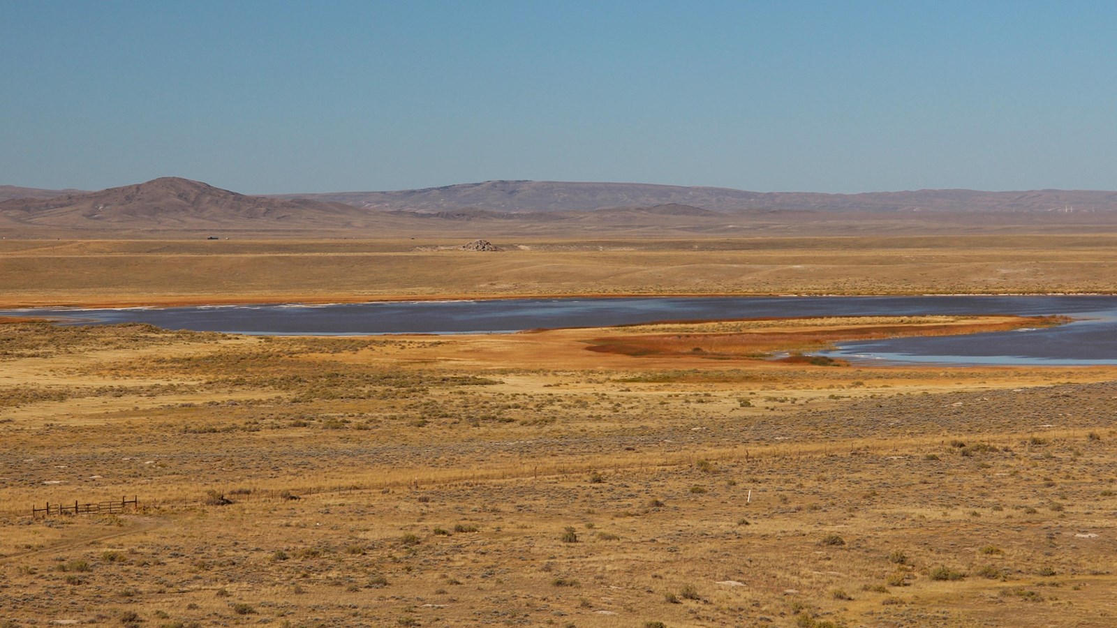 View from Independence Rock: brown-orange plain, shrubs, wagon trail, blue lake, granite, mountains.