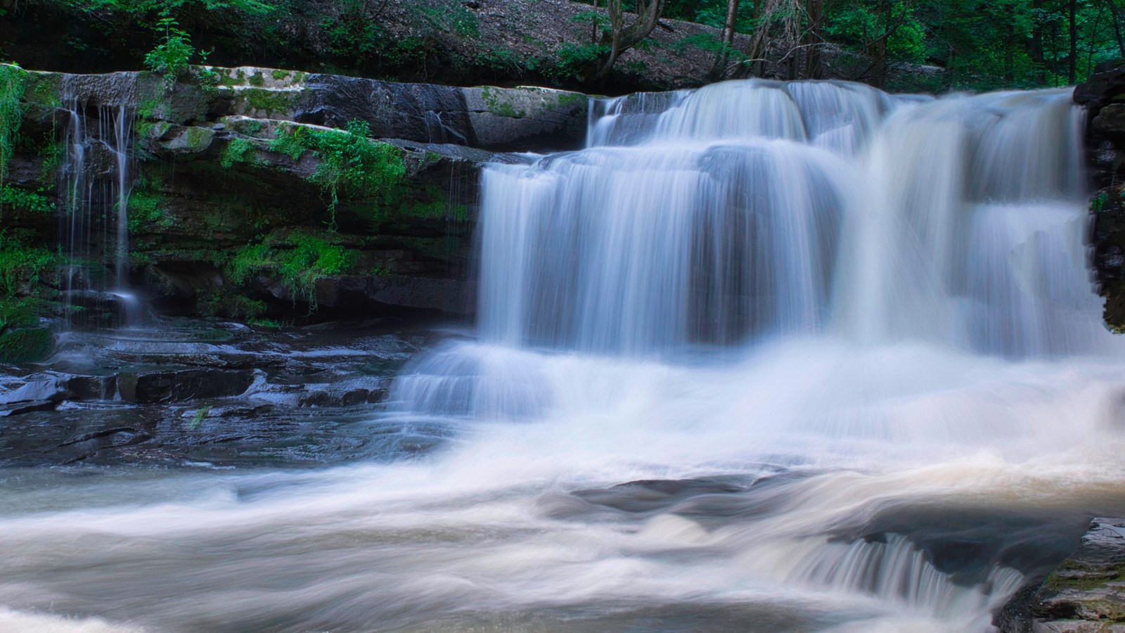 A wide cascade of water flowing down a rocky drop-off into a rock filled pool