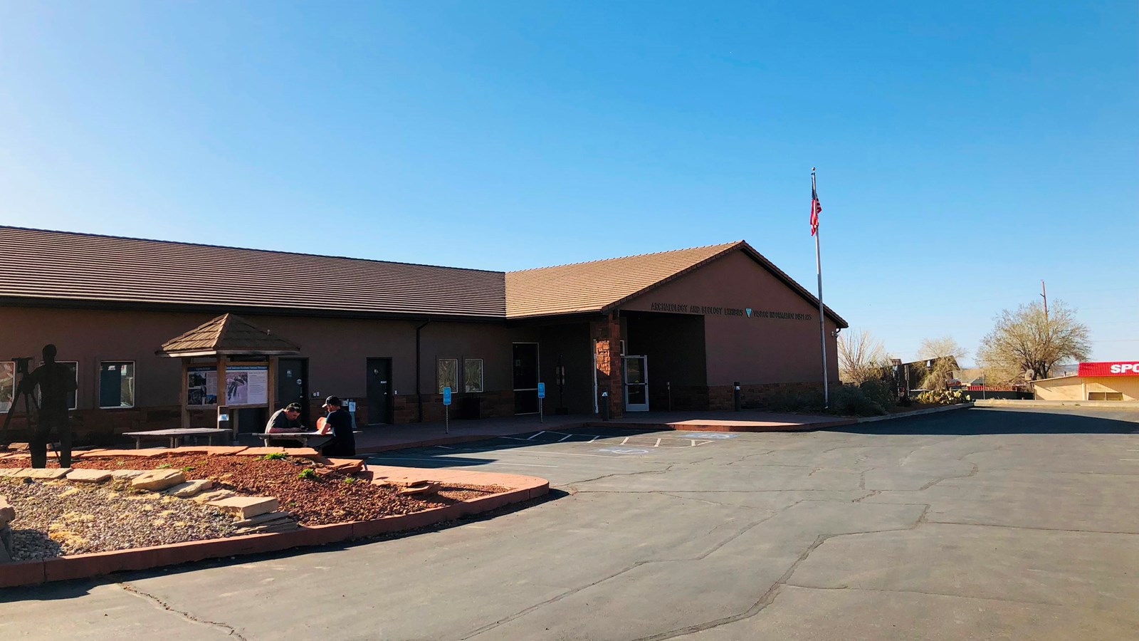 A single story brown building with a dark roof with parking spaces, a gazebo, and flagpole in front.