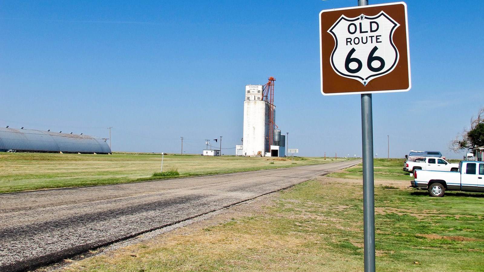 A two-lane paved road surrounded by grass with a concrete building. A sign post reads