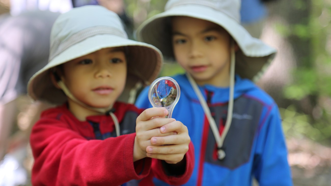 Two children in long sleeved shirts and hats hold up a magnifying lens
