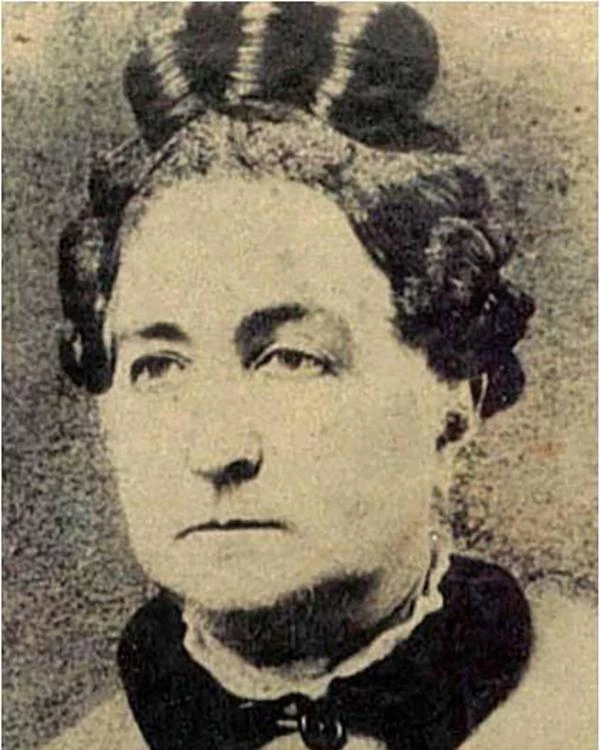A middle aged woman with her hair in a bun and a button up jacket with a black color.