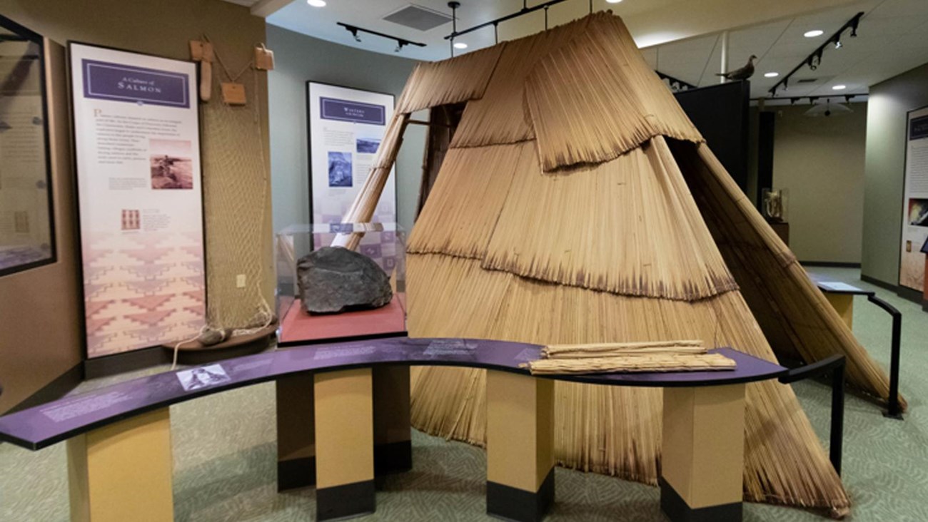 A color photo of an inside room with a windy table. There’s a structure made from organic mats.