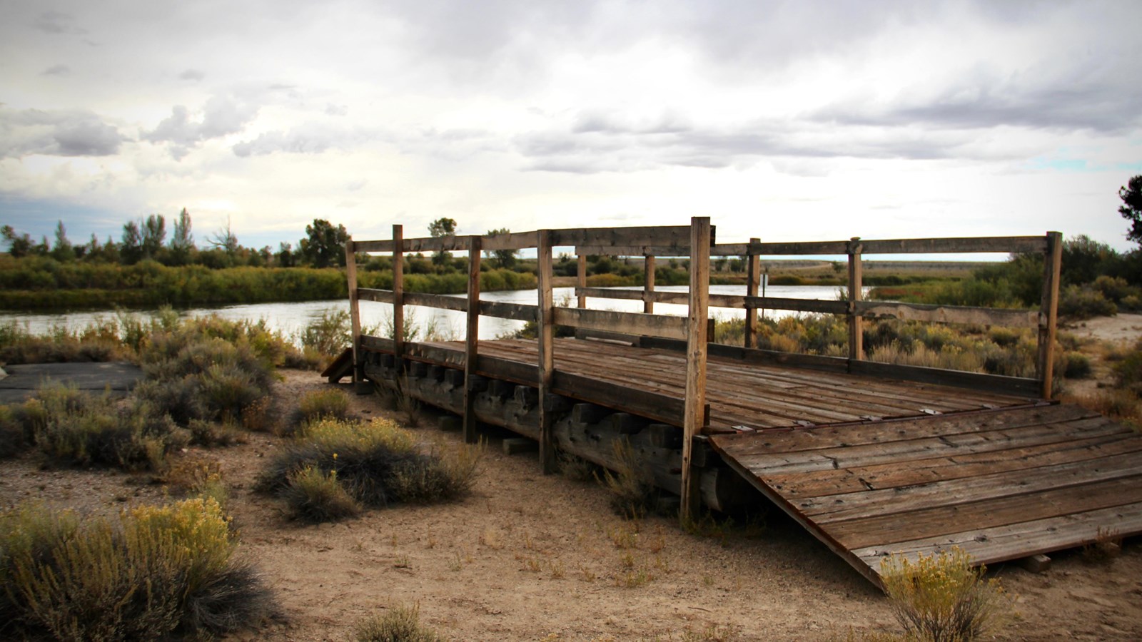 Replica ferry with ramps, posts, and trail; river curves, cottonwoods, and overcast sky.