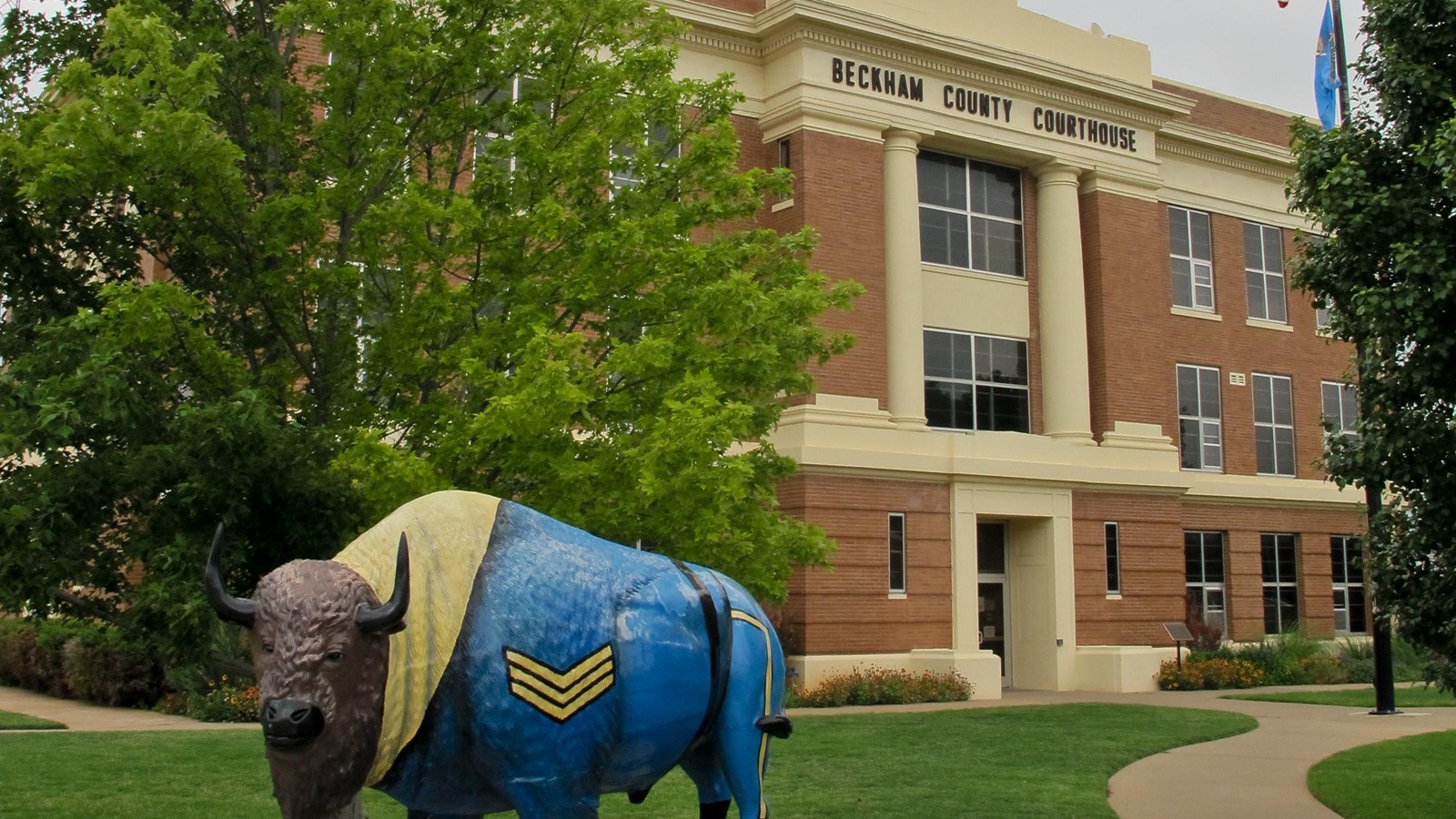 A brown courthouse with white detailing. Out front is a green lawn, tree, and a bison sculpture.