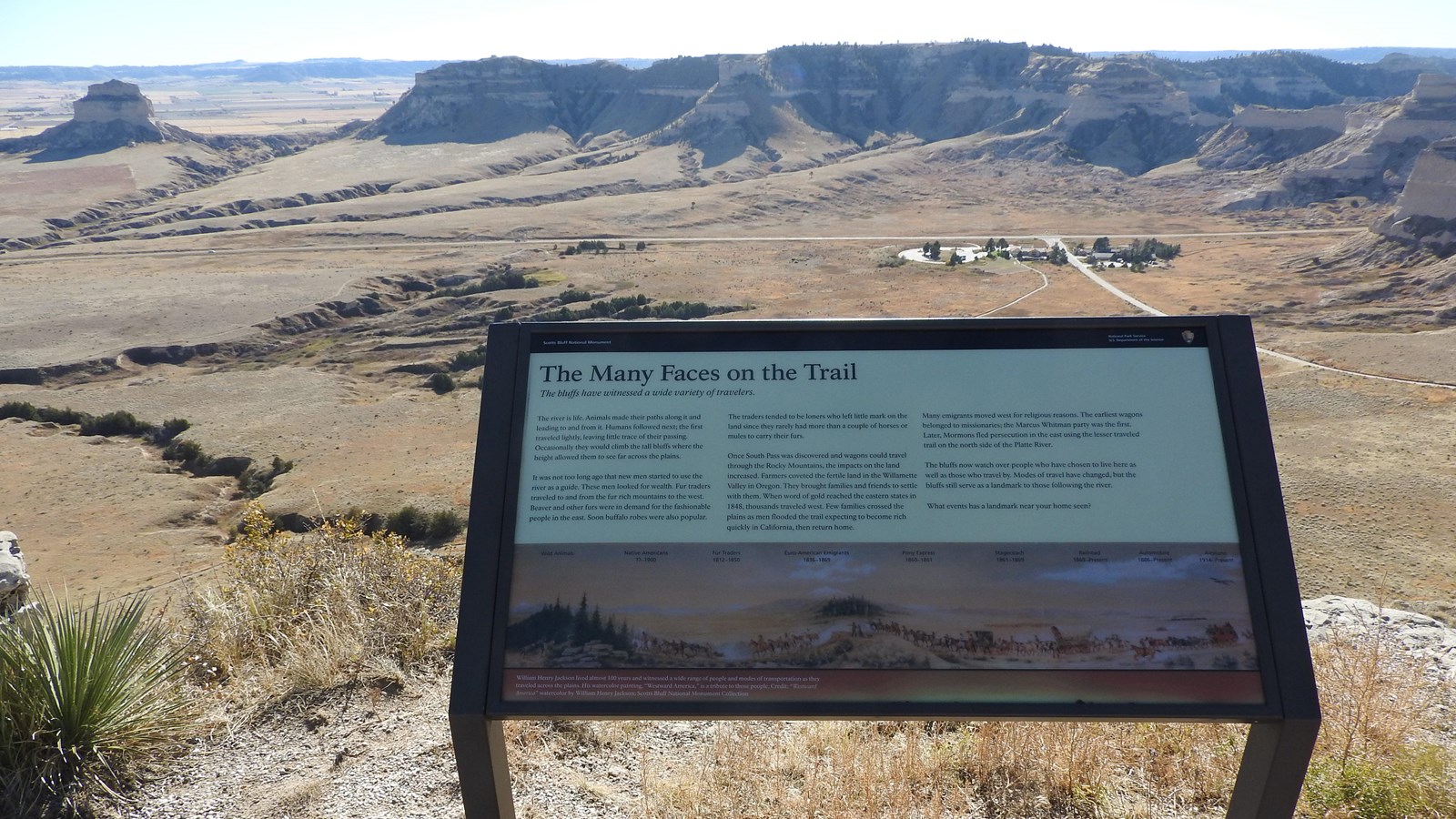 An interpretive wayside panel sits at an overlook of a cluster of buildings and a gap between bluffs