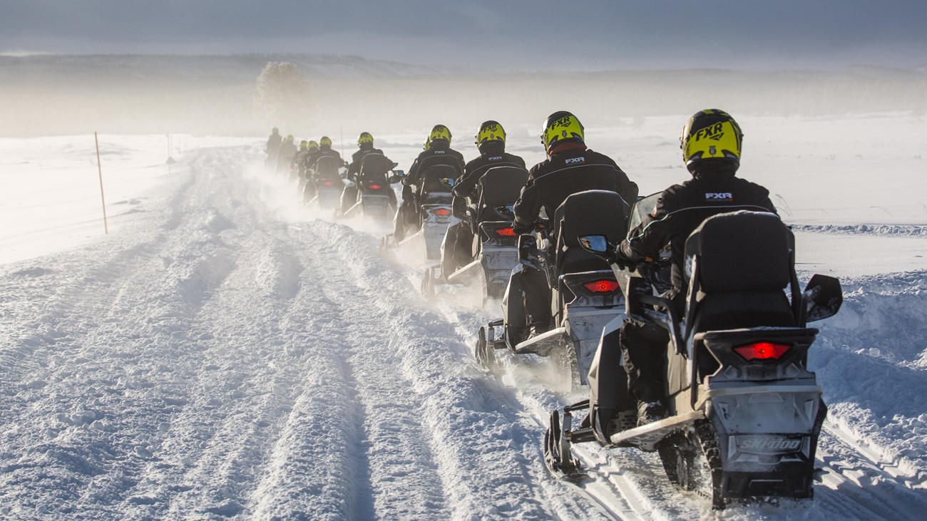A line of snowmobiles move along a snowy track that leads across a snow-covered valley.