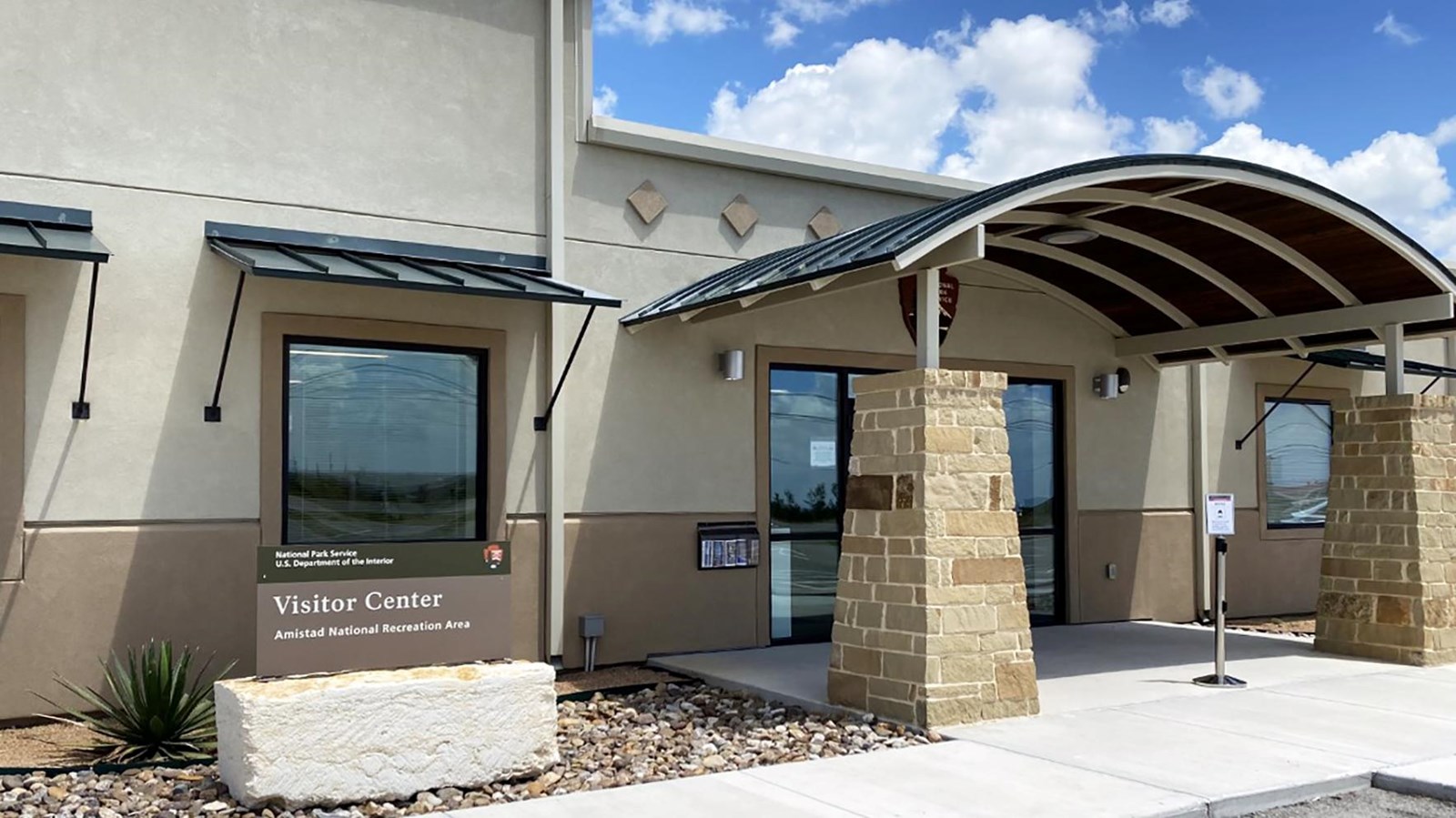 Front of building with arched entrance overhang, small visitor center sign, sidewalk, and glass door