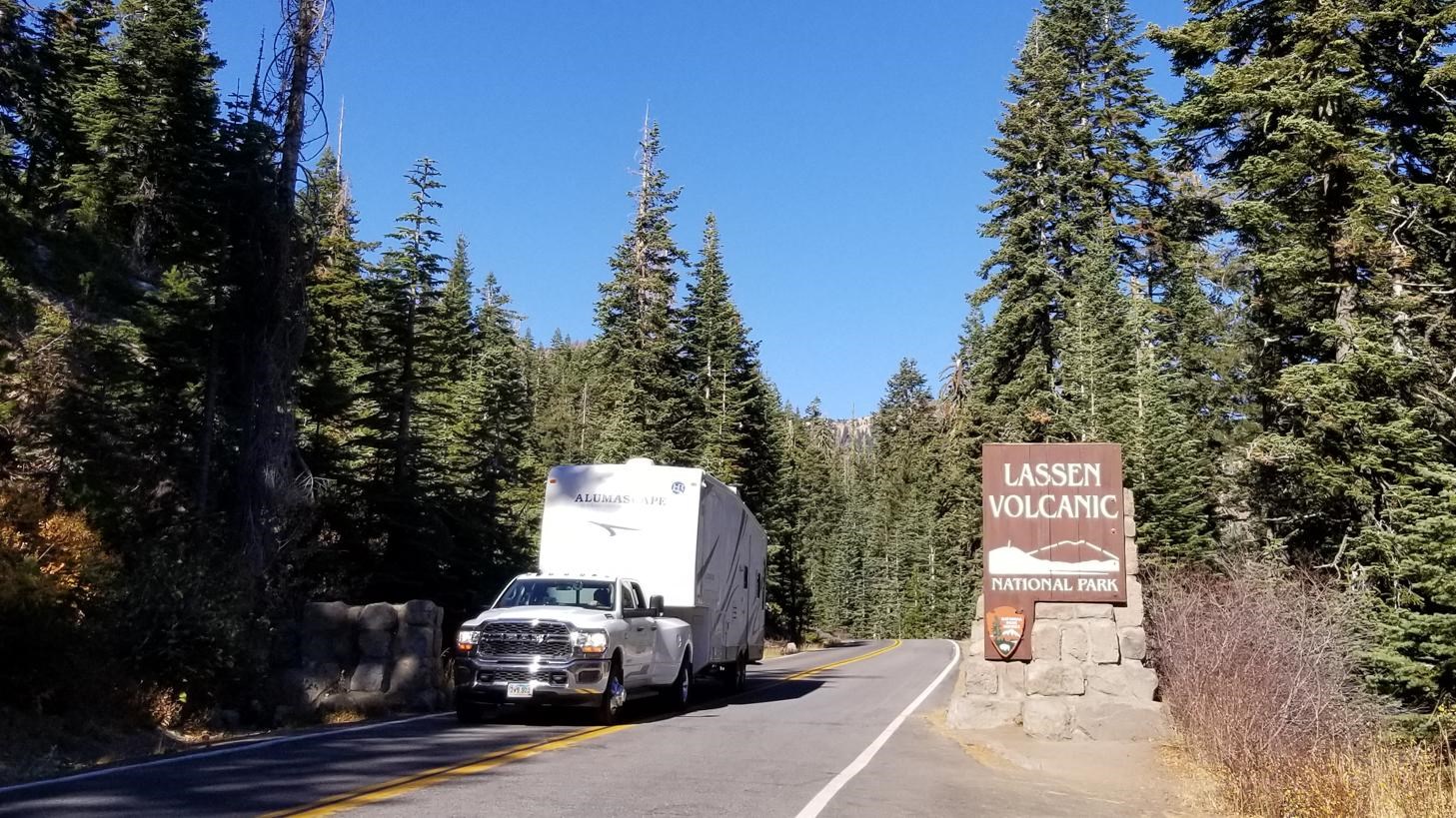 A white truck pulling a camping trailer drives past a wooden sign that reads Lassen Volcanic Nationa