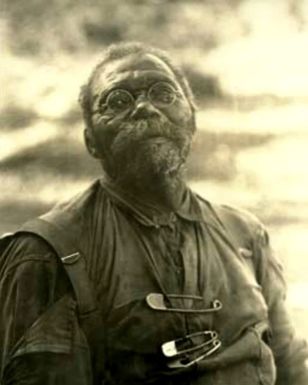 A back and white photo of a man wearing glasses. Large blanket pins are on his shirt.