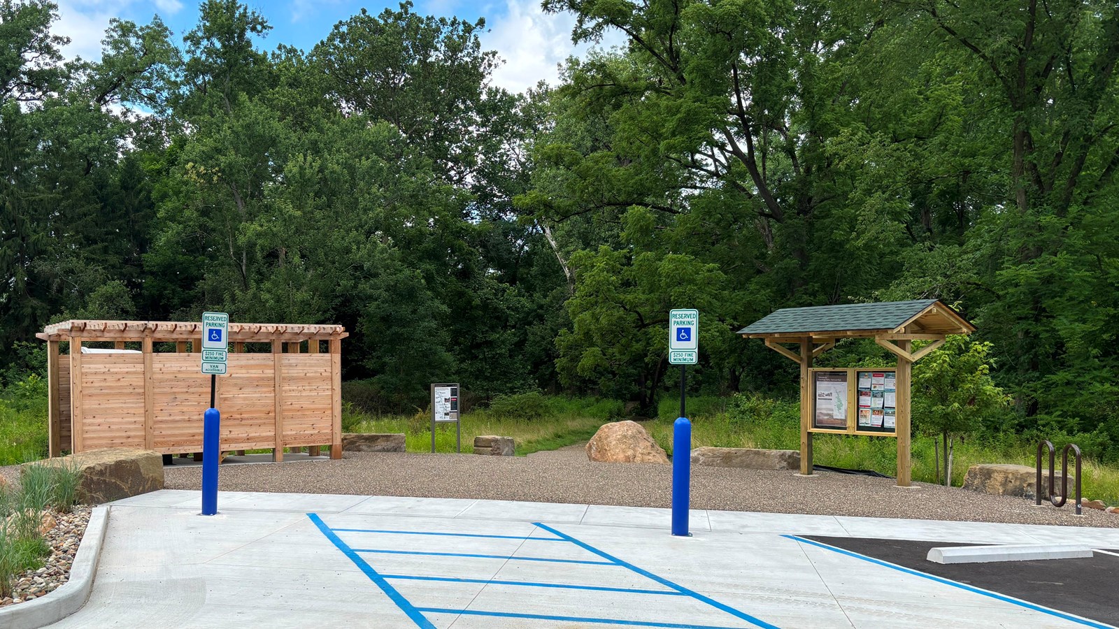 A wooden structure and roofed kiosk near the edge of a parking lot; a trail leads into a forest.