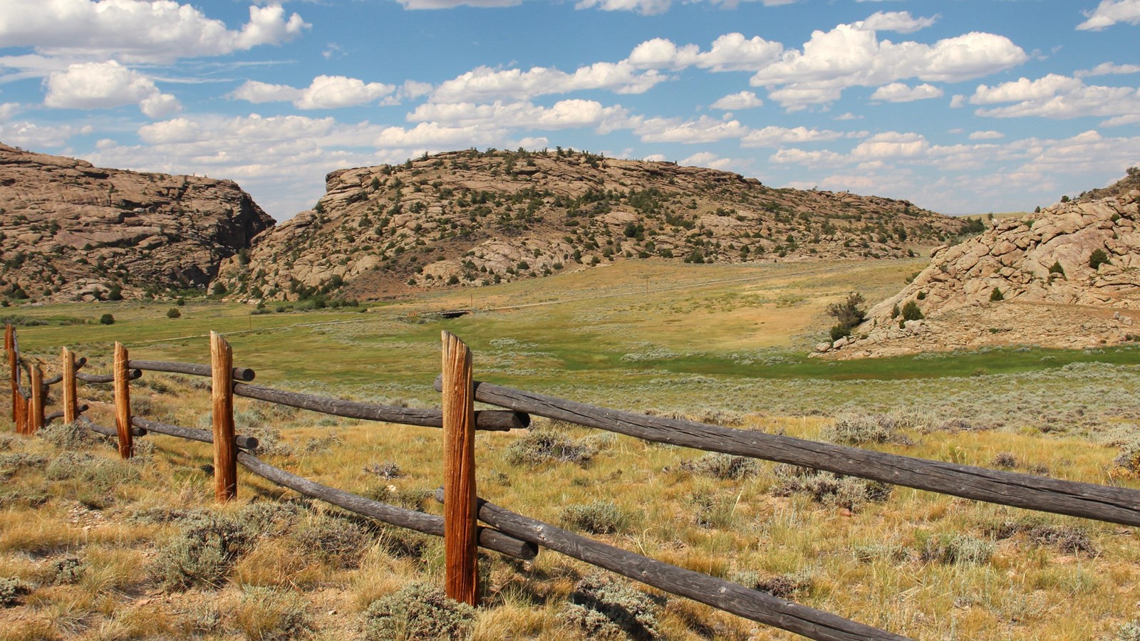 A fence runs along the bottom of the photo with grass, brush, and granite formations under blue sky.