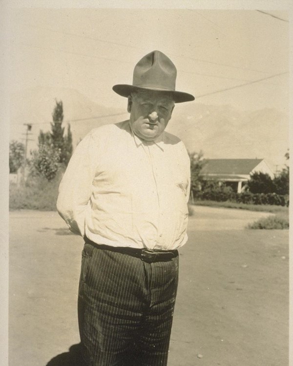 Black and shite photo of man wearing a white bottom-up shirt, slacks, and hat 