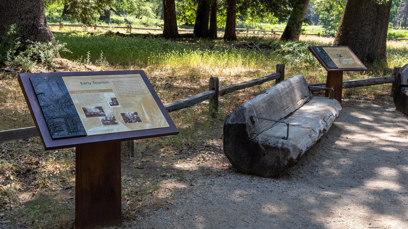 Two wayside exhibits at the edge of a paved path with a wooden tree trunk bench in between them