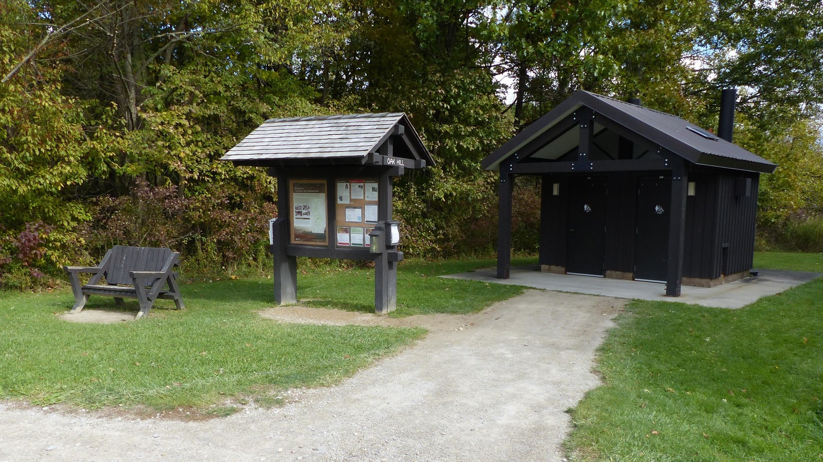 “Oak Hill” kiosk stands between a wooden bench and a two door restroom with trees in the background.