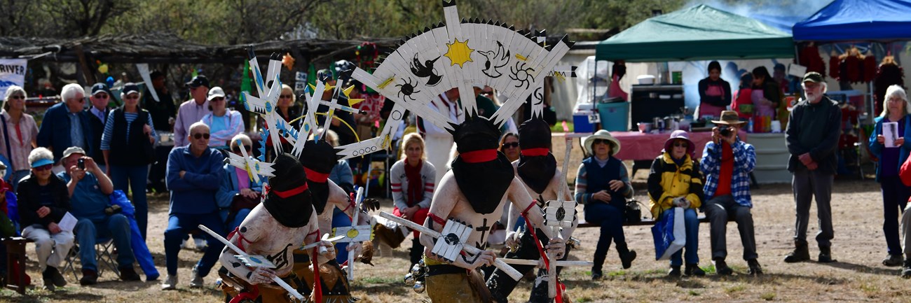 apache crown dancers with large head pieces dance with audience watching and church in background