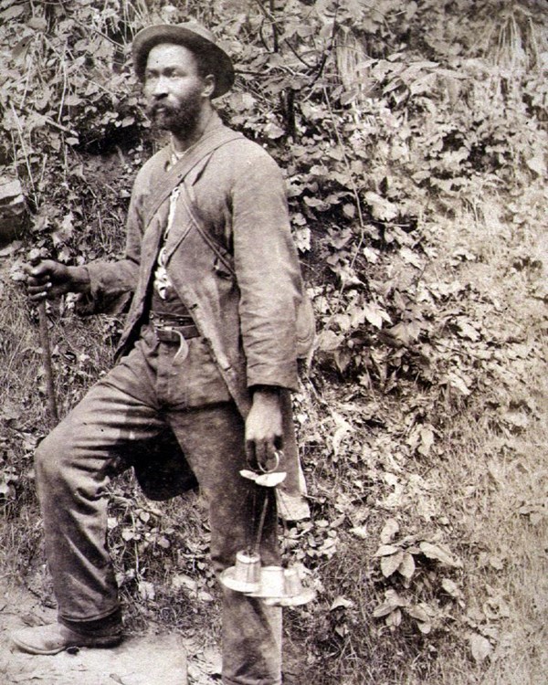 A black and white photo of a man standing on stairs near a cave entrance. He is holding a lantern.