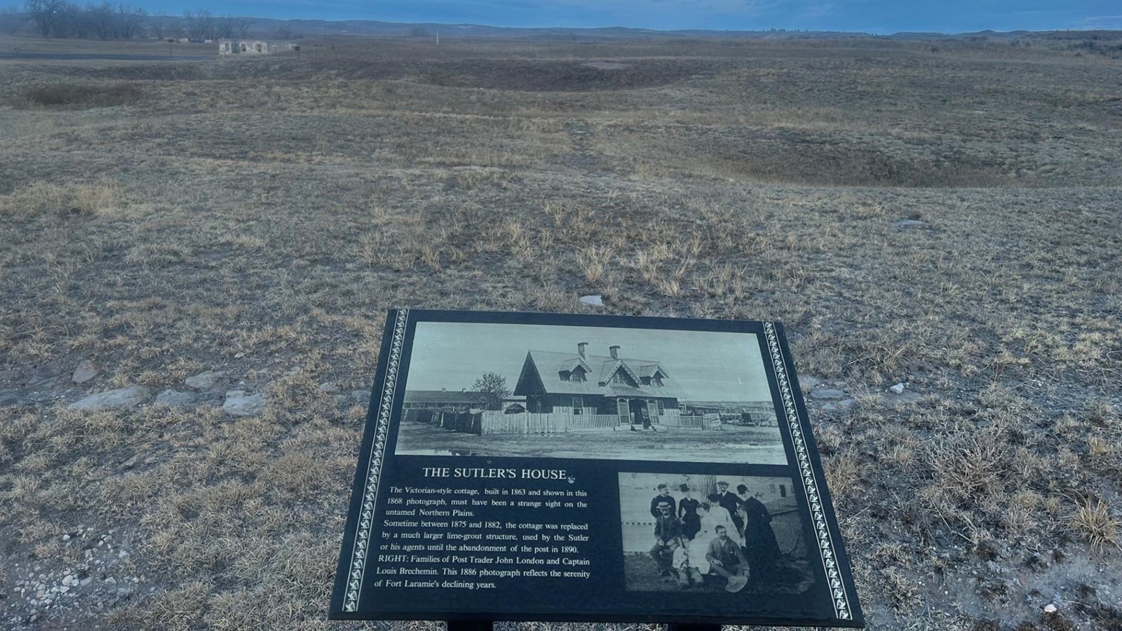 Historical plaque in a dry prairie shows The Sutler\'s House; distant ruins and a cloudy blue sky.