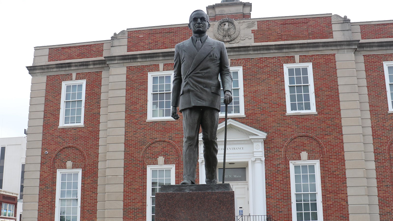 A bronze statue of Harry S Truman wearing a suit, glasses, and walking with a cane.