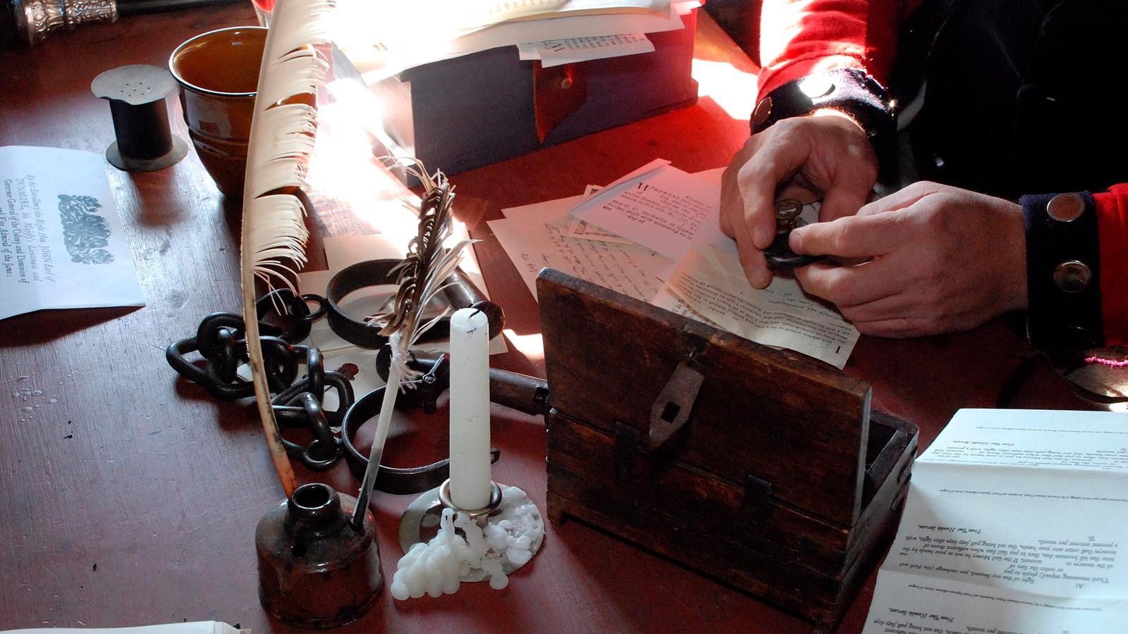 On a table: a candle, papers, and ink. A man\'s hands hold a feather quill.