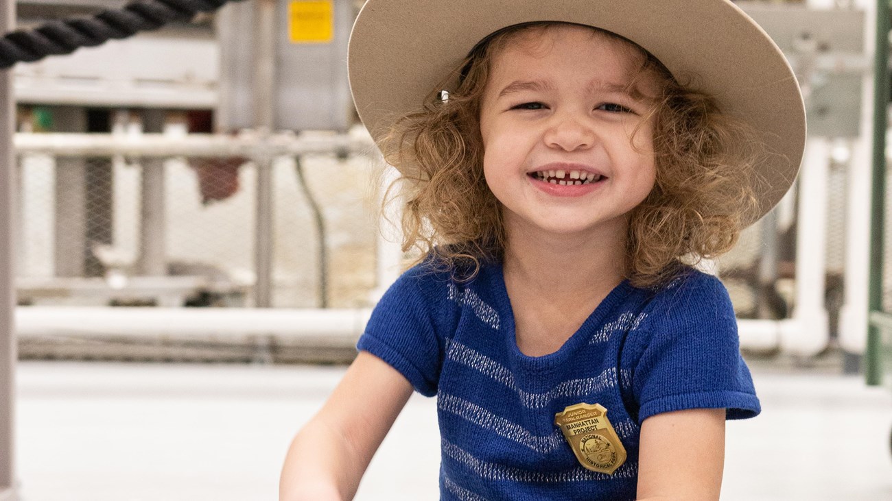 A young girl smiling for the camera, wearing a ranger hat and Junior Ranger badge.