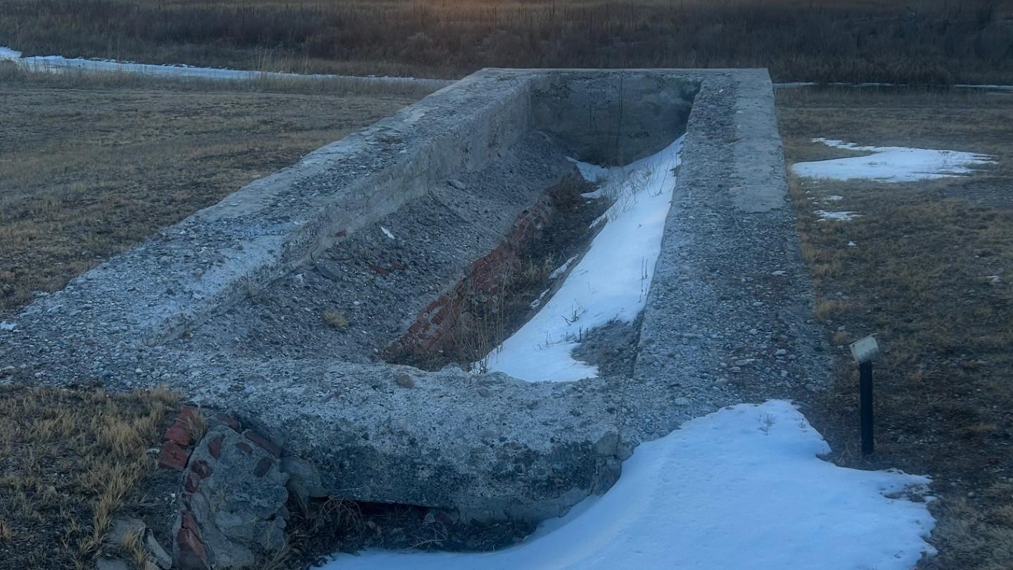 Long rectangular concrete trough in an open field, partly filled with snow, with leafless trees.