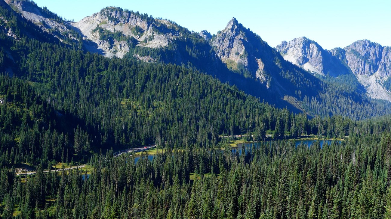 A road curves through a forested valley next to a lake and under a mountain range.
