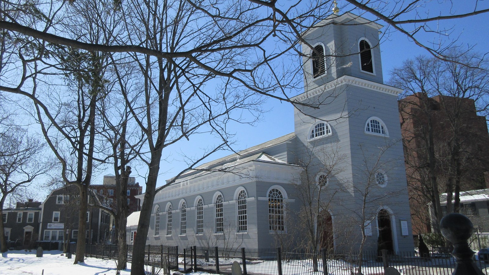 Gray wooden church building with short square steeple at front, and large arched windows along sides