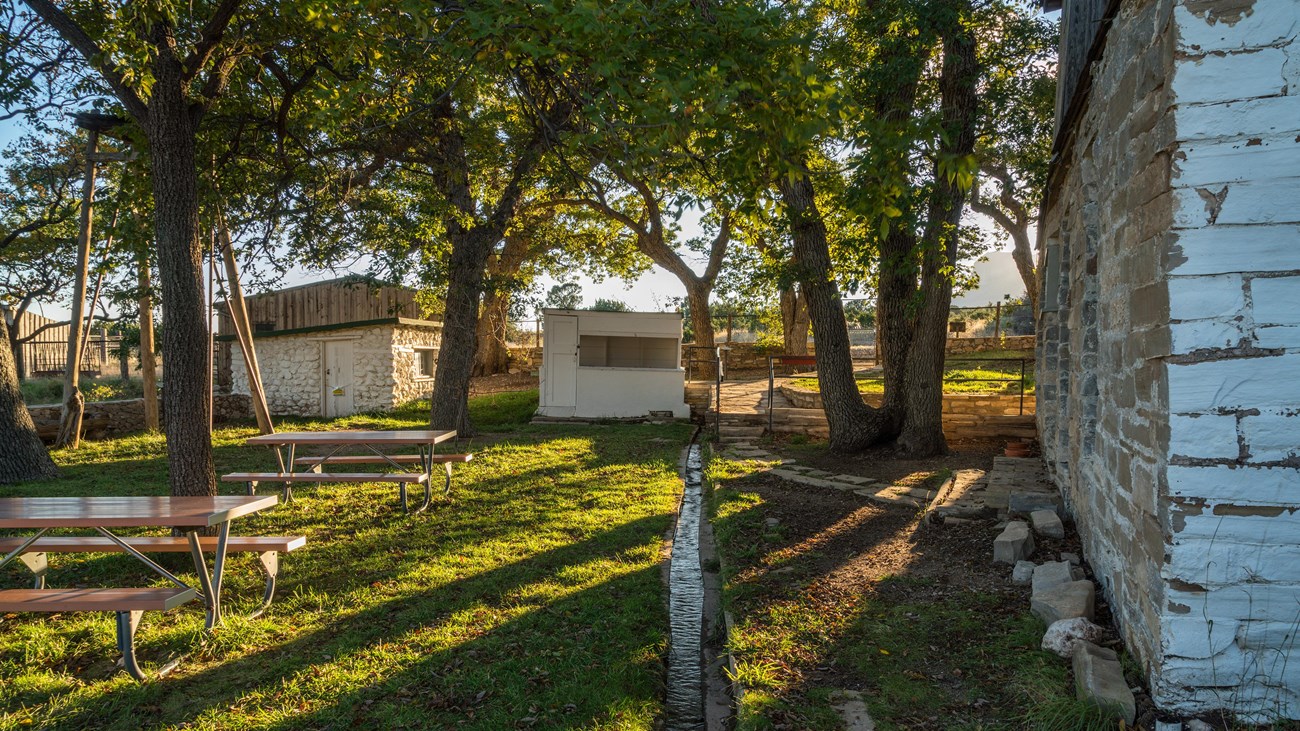 The setting sun casts light and shadows over buildings and a fenced yard with trees.