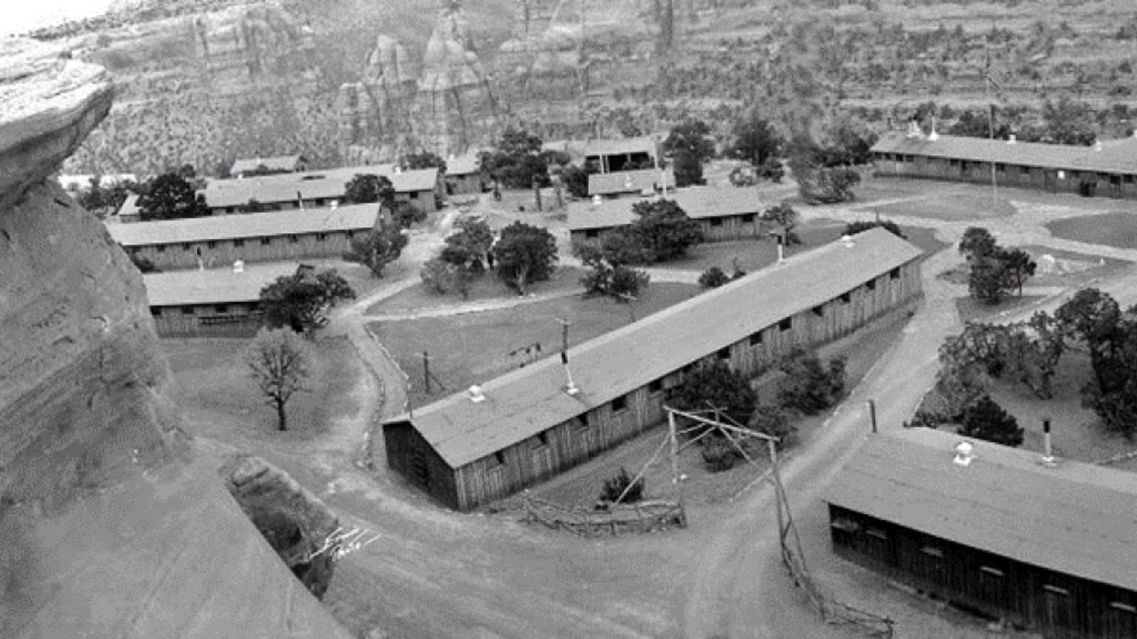 Looking down at 9 long buildings with dirt roads and paths between them. A few trees are nearby.