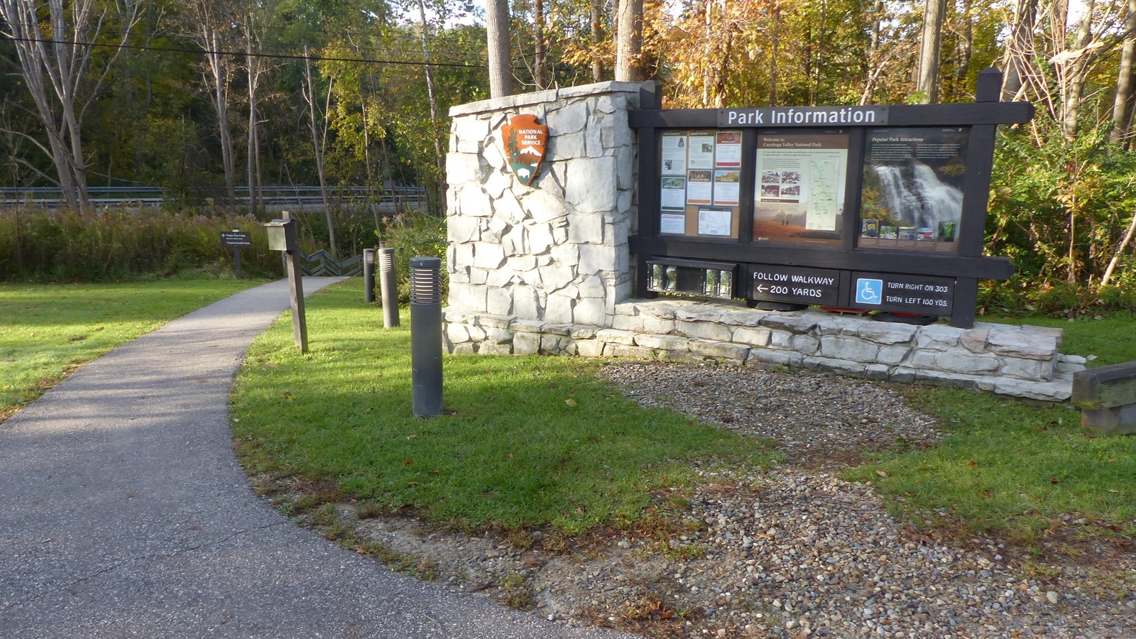 Paved path leads past a three part wooden sign on a large stone base with an NPS arrowhead logo.